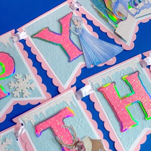 Może przedstawiać: Dekoracyjny baner z r&oacute;żowymi, brokatowymi literami, kt&oacute;re układają się w napis "HAPPY BIRTHDAY". Litery umieszczone są na jasnoniebieskich banerach z r&oacute;żowymi, falistymi obramowaniami. Projekt zawiera wizerunki księżniczki, konia i renifera.