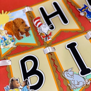 Könnte beinhalten: Ein farbenfrohes Banner mit den Buchstaben "BIRTHDAY" in Schwarz und Weiß, mit Illustrationen von Zeichentrickfiguren. Das Banner besteht aus beigefarbenen Wimpeln mit einem goldfarbenen, gewellten Rand und weißen Bändern. Der Hintergrund ist rot.