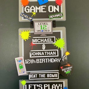 May include: A black and grey game-themed birthday party sign with the text "GAME ON", "LEVEL UP", "LEVEL 12 UNLOCKED", "ADVANCE", "MICHAEL & JOHNATHAN 12th BIRTHDAY", "BEAT THE BOMB", "LET'S PLAY!" and "BIRTHDAY BLAST!" The sign is decorated with colourful paint splatters and a blue lock.
