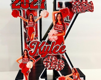Può includere: Una grande lettera K decorativa tridimensionale in nero, bianco e rosso. Presenta il testo 2027, Senior e il nome Kylee. La lettera è decorata con diversi ritagli fotografici di una cheerleader in uniforme rossa con pon pon.