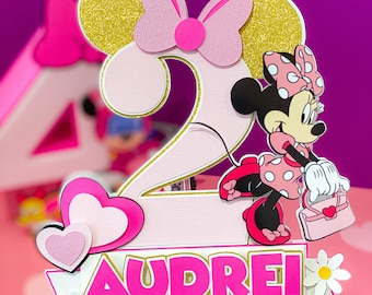 Könnte beinhalten: Rosa-goldener Minnie Mouse-Kuchentopper mit der Zahl 2 und dem Namen Audrei. Der Topper hat eine rosa Schleife, weiße Punkte und eine rosa Handtasche.