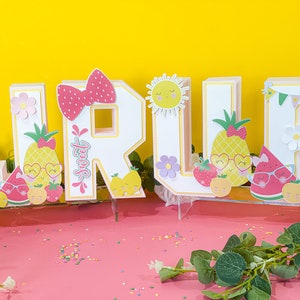 May include: A set of colorful, decorated letters spelling out the name "JRUE" with a summer fruit theme. The letters are made of cardboard and have a white background with a pink outline. Each letter is decorated with various fruits, flowers, and other summer-themed elements. The letters are sitting on a pink surface with sprinkles.