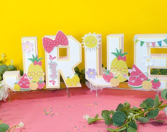 May include: A set of colorful, decorated letters spelling out the name "JRUE" with a summer fruit theme. The letters are made of cardboard and have a white background with a pink outline. Each letter is decorated with various fruits, flowers, and other summer-themed elements. The letters are sitting on a pink surface with sprinkles.