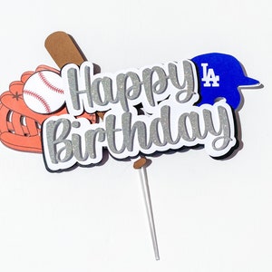 May include: Silver glitter "Happy Birthday" cake topper with a baseball glove, baseball, bat, and a blue baseball cap with "LA" on it.