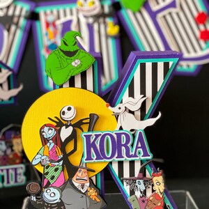 K&ouml;nnte beinhalten: Ein Kuchentopper in Form eines gestreiften Buchstabens K in Lila, Schwarz und Wei&szlig; mit einem gelben Kreis dahinter. Der Kreis zeigt Figuren aus dem Film Nightmare Before Christmas, darunter Jack Skellington, Sally und Oogie Boogie. Der Buchstabe K zeigt auch einen wei&szlig;en Geist und den Namen "KORA" in lila Buchstaben.