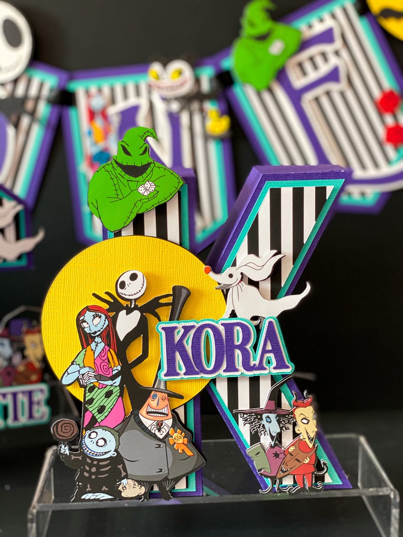 K&ouml;nnte beinhalten: Ein Kuchentopper in Form eines gestreiften Buchstabens K in Lila, Schwarz und Wei&szlig; mit einem gelben Kreis dahinter. Der Kreis zeigt Figuren aus dem Film Nightmare Before Christmas, darunter Jack Skellington, Sally und Oogie Boogie. Der Buchstabe K zeigt auch einen wei&szlig;en Geist und den Namen "KORA" in lila Buchstaben.