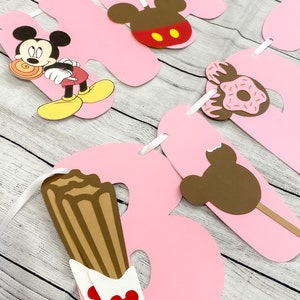 Könnte beinhalten: Rosa Banner mit Mickey Mouse-Charakteren und Lebensmitteln. Das Banner enthält die Buchstaben "A", "B" und "Y" mit Mickey Mouse-Ohren, eine Mickey Mouse, die einen Teller hält, einen Donut, einen Churro und einen Mickey Mouse-Eis am Stiel.