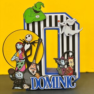 Puede incluir: Un adorno para pastel a rayas blancas y negras con un borde azul. El adorno presenta personajes de la pel&iacute;cula Pesadilla antes de Navidad, incluyendo a Jack Skellington, Sally, Oogie Boogie y Zero. El adorno tambi&eacute;n incluye el nombre "DOMINIC" en letras blancas.