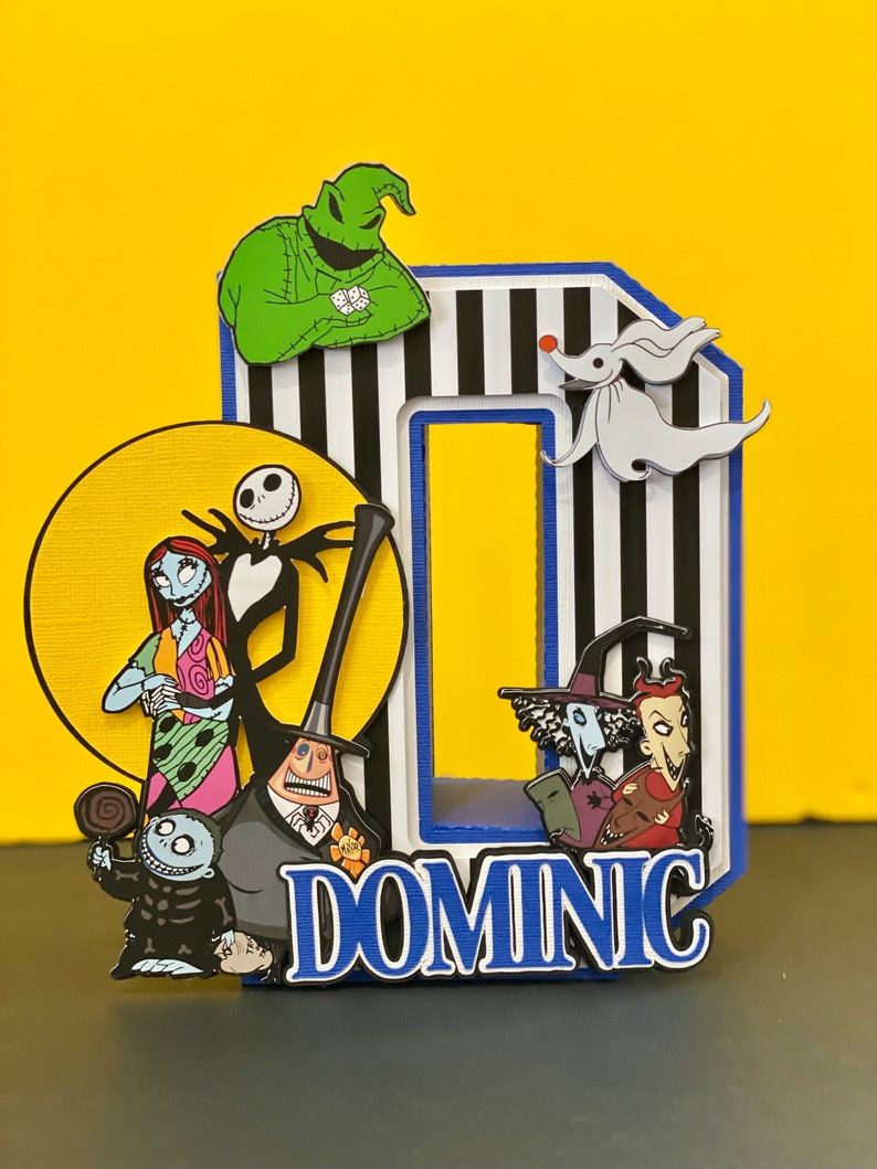 Puede incluir: Un adorno para pastel a rayas blancas y negras con un borde azul. El adorno presenta personajes de la pel&iacute;cula Pesadilla antes de Navidad, incluyendo a Jack Skellington, Sally, Oogie Boogie y Zero. El adorno tambi&eacute;n incluye el nombre "DOMINIC" en letras blancas.