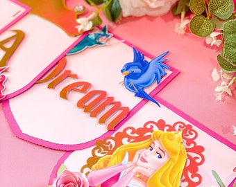 Peut inclure: Une bannière rose et blanche avec le mot "Dream" en lettres dorées. La bannière représente une princesse avec une couronne, un oiseau bleu et des roses roses.