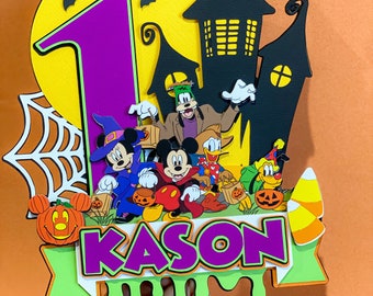 May include: A Halloween-themed cake topper featuring Mickey Mouse, Goofy, Donald Duck, and Pluto dressed in costumes. The topper includes a purple number one, a black haunted house, a yellow moon, and a green banner with the name "KASON" in purple letters.