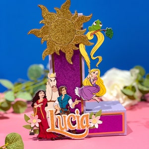 May include: A purple and gold themed cake topper featuring characters from the Disney movie Tangled. The topper includes Rapunzel, Flynn Rider, Pascal the chameleon, and Maximus the horse. The topper also includes a gold sun and the name "Lucia" in gold glitter lettering.