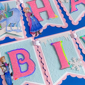 Może przedstawiać: Świąteczny baner z r&oacute;żowymi, brokatowymi literami, kt&oacute;re układają się w napis "HAPPY BIRTHDAY". Litery znajdują się na jasnoniebieskich proporczykach ze wzorem płatka śniegu i r&oacute;żowymi, falistymi krawędziami. Wycięcia postaci i biała wstążka dopełniają projekt.