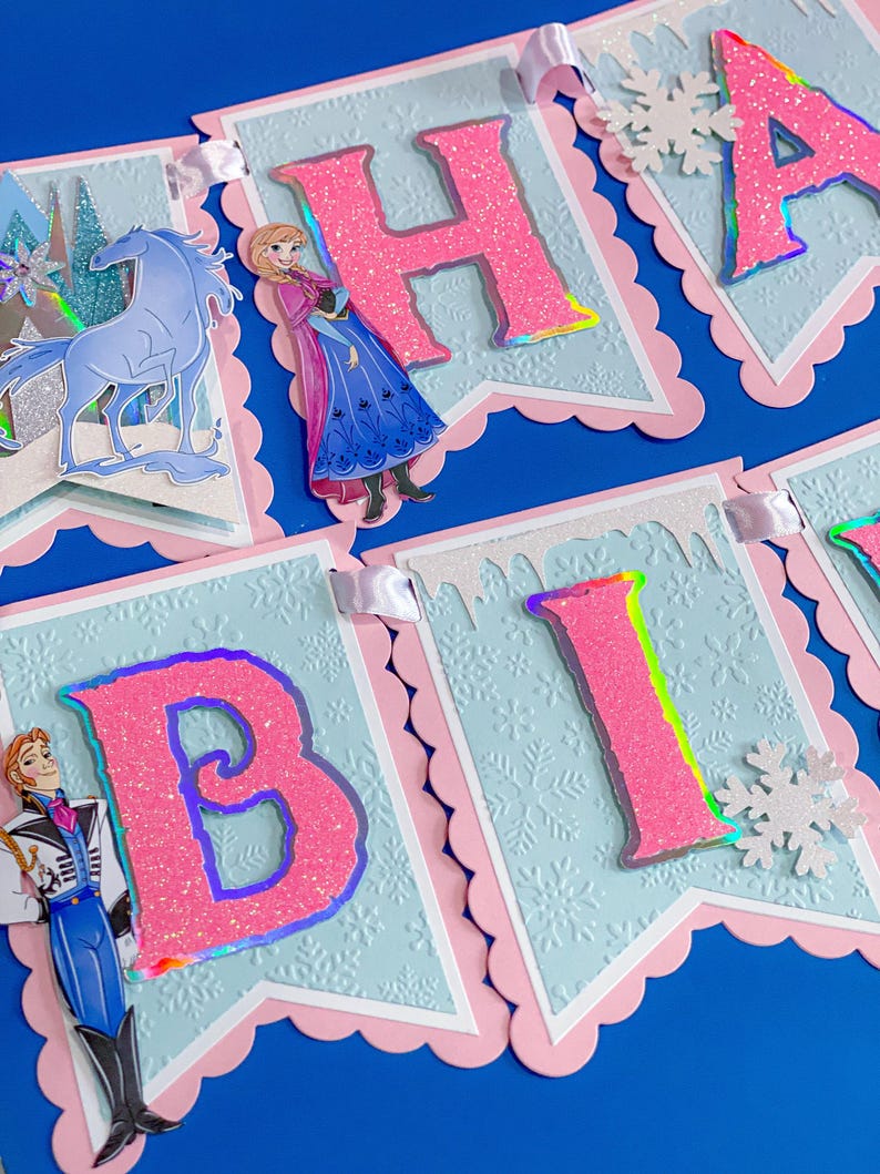 Może przedstawiać: Świąteczny baner z r&oacute;żowymi, brokatowymi literami, kt&oacute;re układają się w napis "HAPPY BIRTHDAY". Litery znajdują się na jasnoniebieskich proporczykach ze wzorem płatka śniegu i r&oacute;żowymi, falistymi krawędziami. Wycięcia postaci i biała wstążka dopełniają projekt.