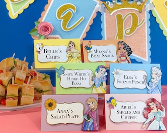 May include: Colorful table setting with themed food labels featuring Disney characters. Labels include "Belle's Chips," "Moana's Boat Snack," "Snow White's Heigh-Ho Pizza," "Elsa's Frozen Punch," "Anna's Salad Plate," and "Ariel's Shells and Cheese."