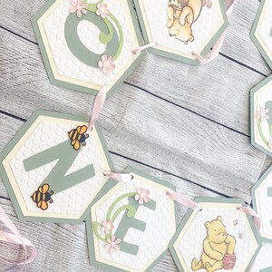 May include: A Winnie the Pooh themed banner for a first birthday party. The banner is made of green and white hexagon-shaped paper with the letters "ONE" and images of Winnie the Pooh holding a honey pot. The banner is strung together with pink ribbon.