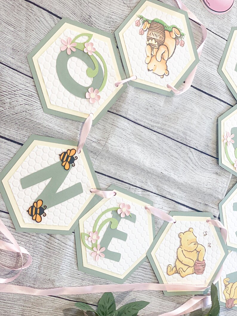 May include: A Winnie the Pooh themed banner for a first birthday party. The banner is made of green and white hexagon-shaped paper with the letters "ONE" and images of Winnie the Pooh holding a honey pot. The banner is strung together with pink ribbon.