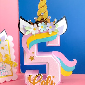 May include: A pink and gold unicorn-themed cake topper shaped like the number 5. The unicorn has a gold horn, a pink mane, and a blue tail. The number 5 is decorated with gold stars and the name "Cali" is written in gold letters on the bottom.