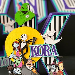 K&ouml;nnte beinhalten: Ein schwarz-wei&szlig; gestreifter Hintergrund mit einem lila und t&uuml;rkisfarbenen Buchstaben K. Der Buchstabe ist mit Figuren aus dem Film Nightmare Before Christmas verziert, darunter Jack Skellington, Sally, Oogie Boogie und Zero. Der Name KORA ist in lila Buchstaben auf dem Buchstaben K geschrieben.