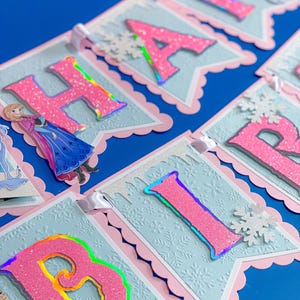 Może przedstawiać: Baner z napisem "HAPPY BIRTHDAY" z r&oacute;żowymi, brokatowymi literami i akcentami w kształcie płatk&oacute;w śniegu. Baner ma jasnoniebieskie tło z r&oacute;żową, falistą obw&oacute;dką. Widoczne są r&oacute;wnież postacie z popularnej animacji.