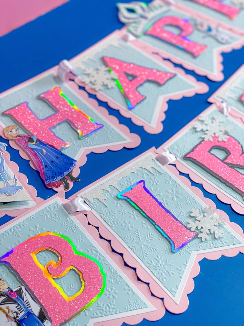 Może przedstawiać: Baner z napisem "HAPPY BIRTHDAY" z r&oacute;żowymi, brokatowymi literami i akcentami w kształcie płatk&oacute;w śniegu. Baner ma jasnoniebieskie tło z r&oacute;żową, falistą obw&oacute;dką. Widoczne są r&oacute;wnież postacie z popularnej animacji.