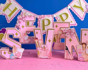 Può includere: Un set di lettere tridimensionali glitterate rosa che compongono la parola SEVEN, decorate con immagini di pennelli da trucco, flaconi di profumo, rossetti e ciprie. Sullo sfondo è appeso uno striscione rosa e oro con la scritta HAPPY.