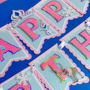 Może przedstawiać: Baner z napisem "HAPPY" z r&oacute;żowymi, brokatowymi literami, płatkami śniegu i wycięciami postaci. Baner ma jasnoniebieskie tło z r&oacute;żową, falistą obw&oacute;dką. Na początku znajduje się srebrna tiara.