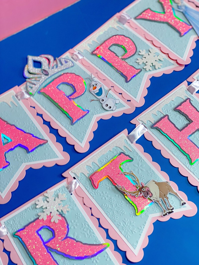 Może przedstawiać: Baner z napisem "HAPPY" z r&oacute;żowymi, brokatowymi literami, płatkami śniegu i wycięciami postaci. Baner ma jasnoniebieskie tło z r&oacute;żową, falistą obw&oacute;dką. Na początku znajduje się srebrna tiara.