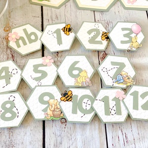 May include: A Winnie the Pooh themed party banner with green and white honeycomb-shaped decorations featuring numbers 1 through 12, a bee, and Winnie the Pooh characters. The banner also includes a "nb" decoration.
