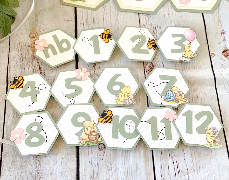 May include: A Winnie the Pooh themed party banner with green and white honeycomb-shaped decorations featuring numbers 1 through 12, a bee, and Winnie the Pooh characters. The banner also includes a "nb" decoration.