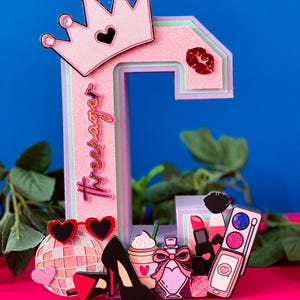 Puede incluir: Letra C rosa decorada con una corona, l&aacute;piz labial, frasco de perfume, tacones altos, una bola de discoteca y una paleta de maquillaje. La letra est&aacute; decorada con purpurina y la palabra "Tres Swag" est&aacute; escrita en el lateral.