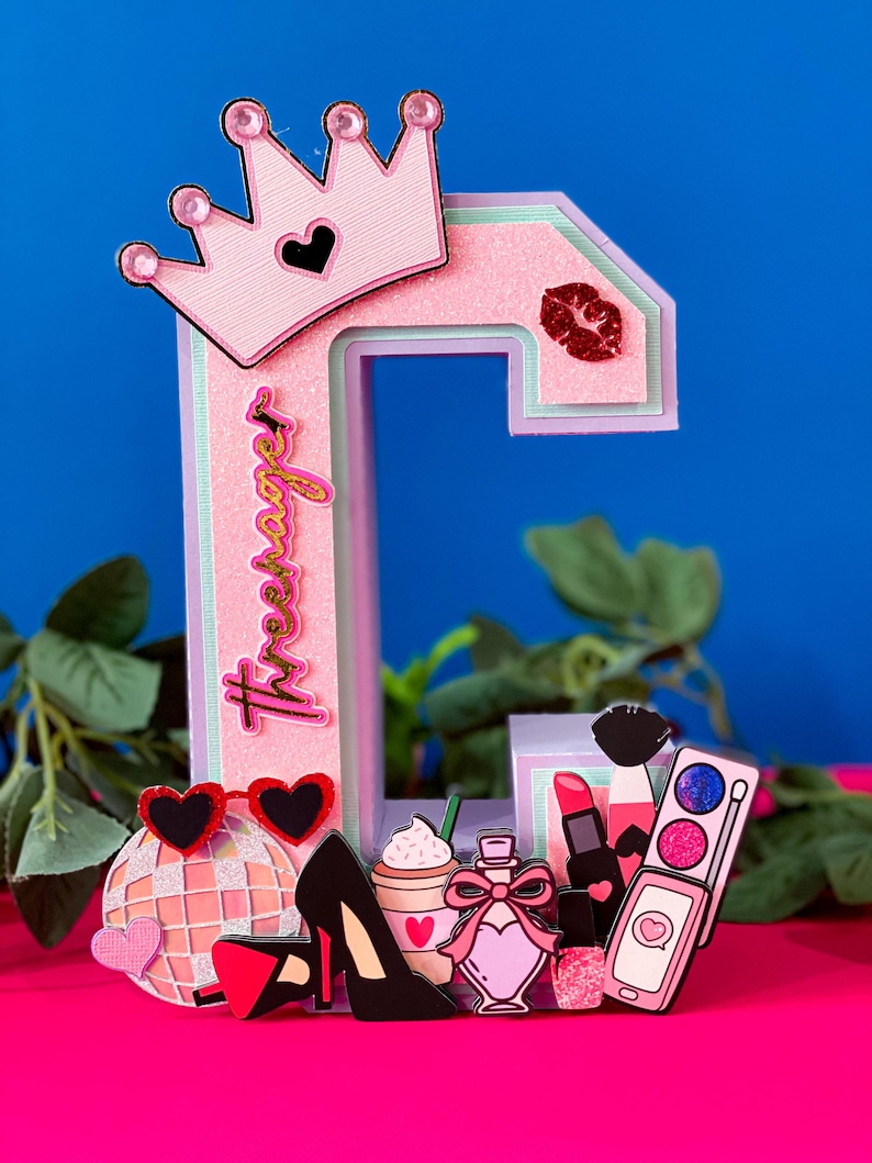 Puede incluir: Letra C rosa decorada con una corona, l&aacute;piz labial, frasco de perfume, tacones altos, una bola de discoteca y una paleta de maquillaje. La letra est&aacute; decorada con purpurina y la palabra "Tres Swag" est&aacute; escrita en el lateral.