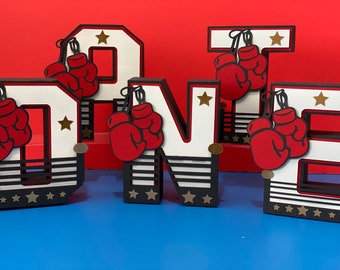 Könnte beinhalten: Weiße Pappbuchstaben, die "ONE" buchstabieren, verziert mit roten Boxhandschuhen und schwarz-weißen Boxring-Designs.