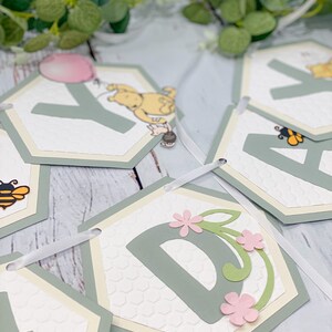 May include: A Winnie the Pooh themed baby shower banner with green and white hexagonal flags. Each flag features a letter of the alphabet, a bee, a flower, or Winnie the Pooh. The flags are connected with a white ribbon.