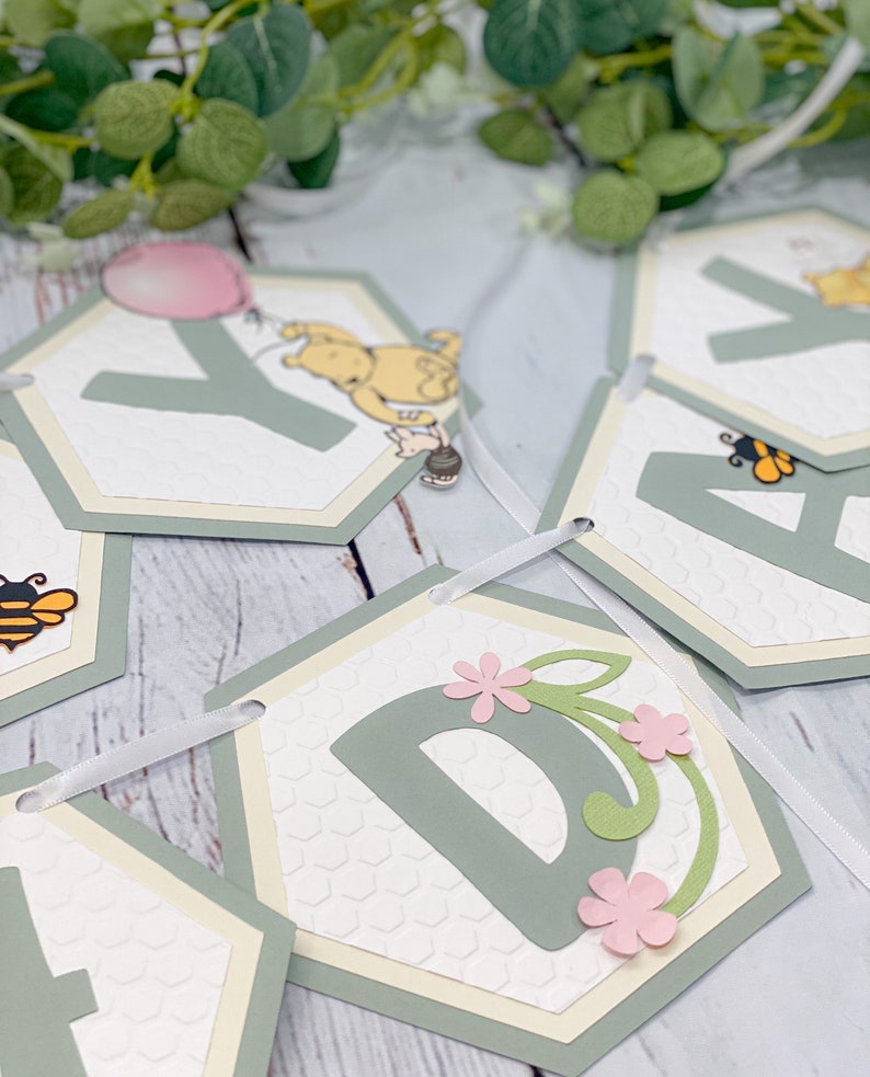 May include: A Winnie the Pooh themed baby shower banner with green and white hexagonal flags. Each flag features a letter of the alphabet, a bee, a flower, or Winnie the Pooh. The flags are connected with a white ribbon.