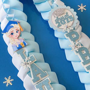 Pode incluir: Um colar de graduação azul claro e branco com um design de floco de neve. O colar tem uma letra "A" de brilho azul e branco e um círculo de brilho azul e branco com o texto "Class of 2023". O colar também tem uma pequena imagem de um personagem de desenho animado com um chapéu de graduação azul.