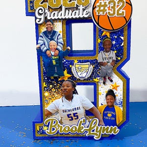 May include: A large, blue, three-dimensional letter B display featuring gold glitter accents, photos of a student, and the text 2026 Graduate, #32, and Brook-Lynn. The display includes a basketball graphic, a graduation cap icon, and a school crest.