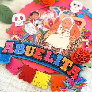 May include: A colorful cake topper featuring the word "Abuelita" in bright letters, a cartoon character in a wheelchair, and a skull with flowers. The topper is decorated with paper flowers, maracas, and a banner.