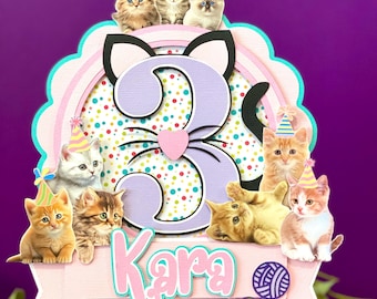 Op de afbeelding: Een paarse en roze 3D-taarttopper met een paars kattenhoofd en het nummer 3 in het midden. De taarttopper heeft een roze banner met de naam "Kara" erop. Er zijn verschillende kleine kittens die feestmutsen dragen rond de taarttopper.