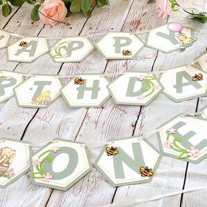 May include: A green and white Winnie the Pooh themed banner with the words "Happy Birthday One" in hexagon-shaped pieces. The banner is decorated with bees, flowers, and Winnie the Pooh characters.