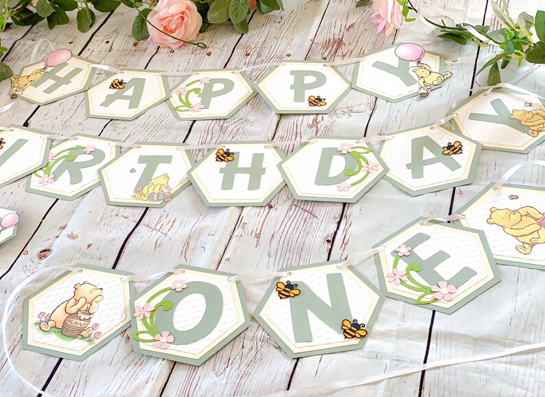 May include: A green and white Winnie the Pooh themed banner with the words "Happy Birthday One" in hexagon-shaped pieces. The banner is decorated with bees, flowers, and Winnie the Pooh characters.