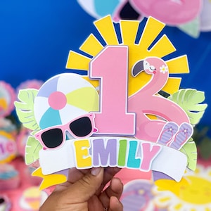May include: A colorful birthday cake topper with a beach theme. It features a pink number 1, a flamingo, sunglasses, a beach ball, and the name "EMILY" on a banner. The background is a bright blue.