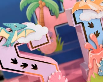 Può includere: Una decorazione tridimensionale del numero 21 nei colori rosa pastello e blu con bordi dorati. Il design include sagome di uno pterodattilo verde, un triceratopo rosa, una palma, un vulcano, nuvole e impronte di dinosauro.