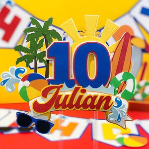 May include: A blue and red cake topper with the number 10 and the name Julian. The topper features a beach theme with palm trees, a beach ball, a surfboard, and a sun.