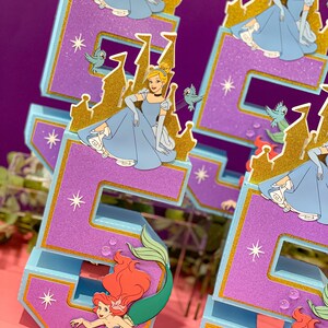 May include: Two blue and purple numbers, 5 and 6, decorated with glitter and gold accents. Each number features a princess character from a Disney movie, Cinderella, sitting on a golden castle. The bottom of each number features a mermaid character, Ariel, with red hair and a green tail.