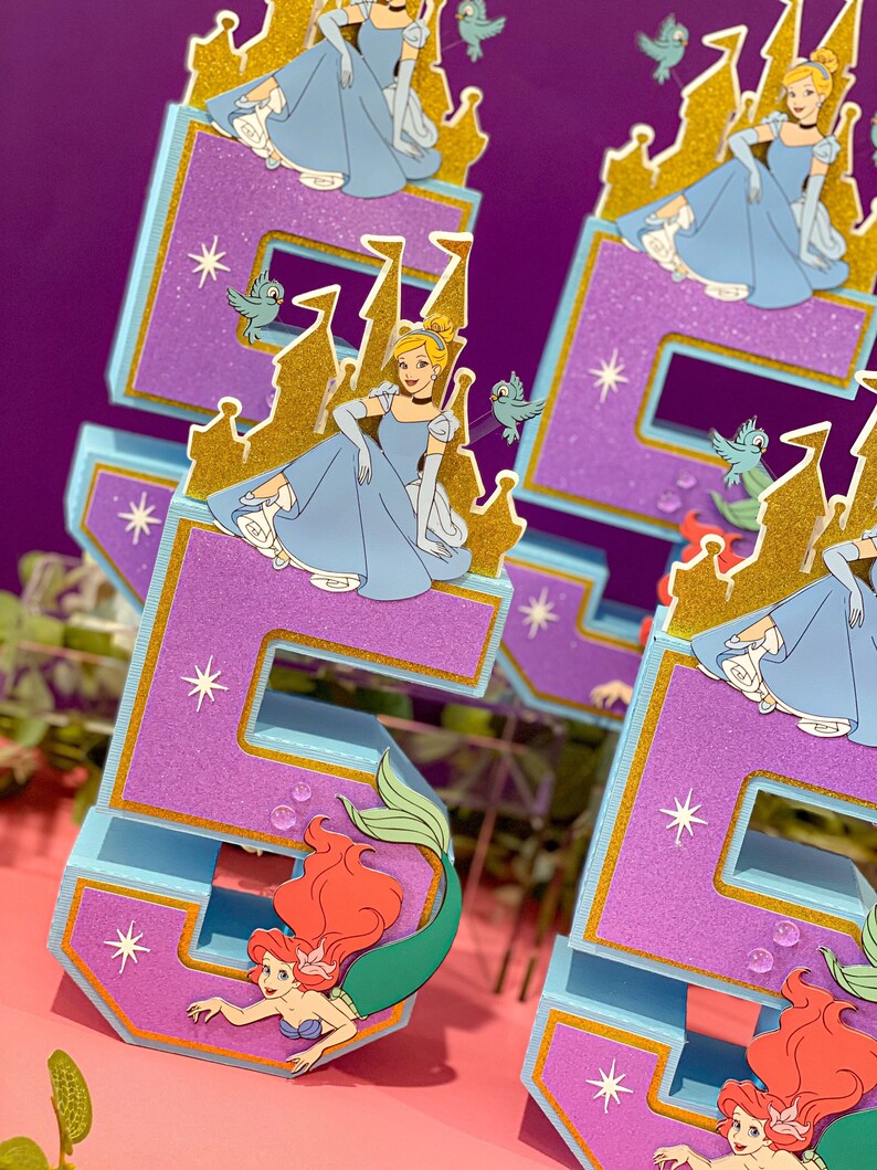 May include: Two blue and purple numbers, 5 and 6, decorated with glitter and gold accents. Each number features a princess character from a Disney movie, Cinderella, sitting on a golden castle. The bottom of each number features a mermaid character, Ariel, with red hair and a green tail.