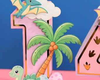 Può includere: Una decorazione tridimensionale a forma di numero uno in rosa chiaro con bordi dorati, raffigurante uno pterosauro verde che vola sopra una palma, un dinosauro verde a terra e due uova decorate, di cui una aperta e rotta.