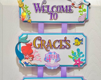 Può includere: Un cartello sospeso a tre livelli con un castello dorato, una sirena e creature marine. Il testo recita Welcome To, Grace's 4th e Birthday. Il cartello è decorato con conchiglie, corallo e un tridente, collegati da nastri di colore viola.