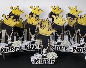 Puede incluir: Un conjunto de seis centros de mesa para fiestas negros y dorados con el número uno, una corona y el nombre "Kiarie". Cada centro de mesa tiene un recipiente de plástico transparente lleno de dinero falso y una cadena dorada.