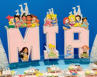 Può includere: Lettere rosa glitter che compongono la parola "MIA" con personaggi delle principesse Disney in cima a ciascuna lettera. Le lettere sono su un supporto bianco con uno sfondo blu.